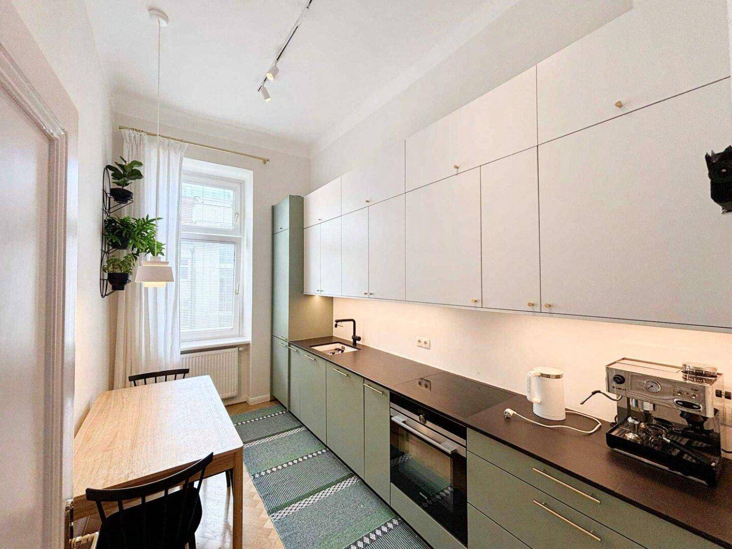 Moderne Küche mit hellgrünen Unterschränken, weißen Oberschränken und einem kleinen Essbereich am Fenster.