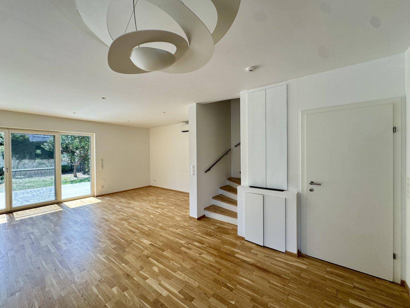 Helles Wohnzimmer mit Parkettboden, moderner Deckenleuchte und Zugang zur Treppe.