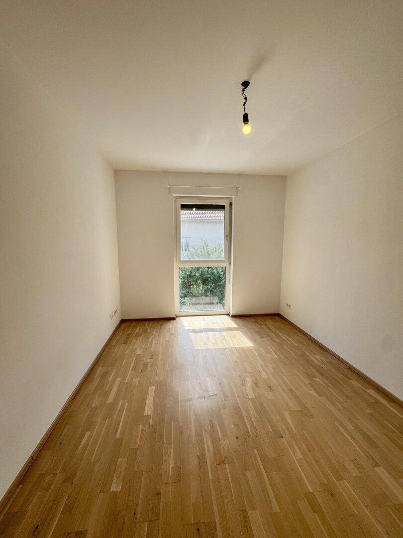 Helles Schlafzimmer mit Parkettboden und Fenster, das viel Sonnenlicht hereinlässt.