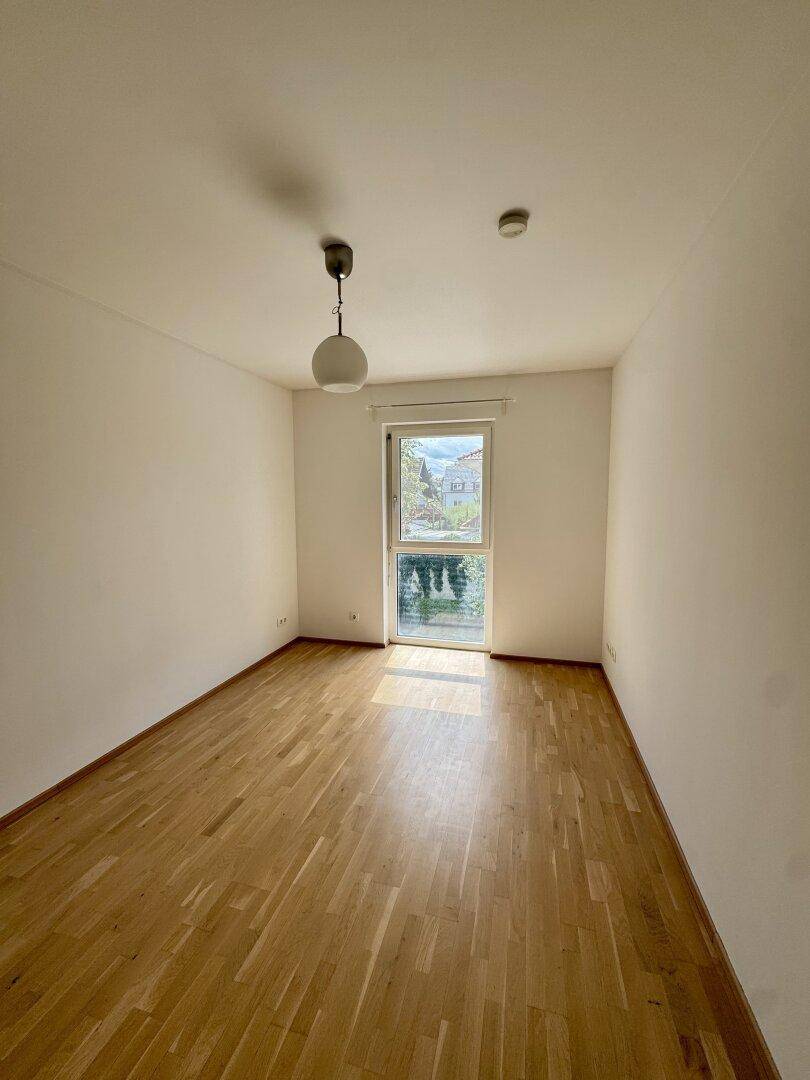 Helles Schlafzimmer mit Parkettboden und großem Fenster, das den Raum erhellt.