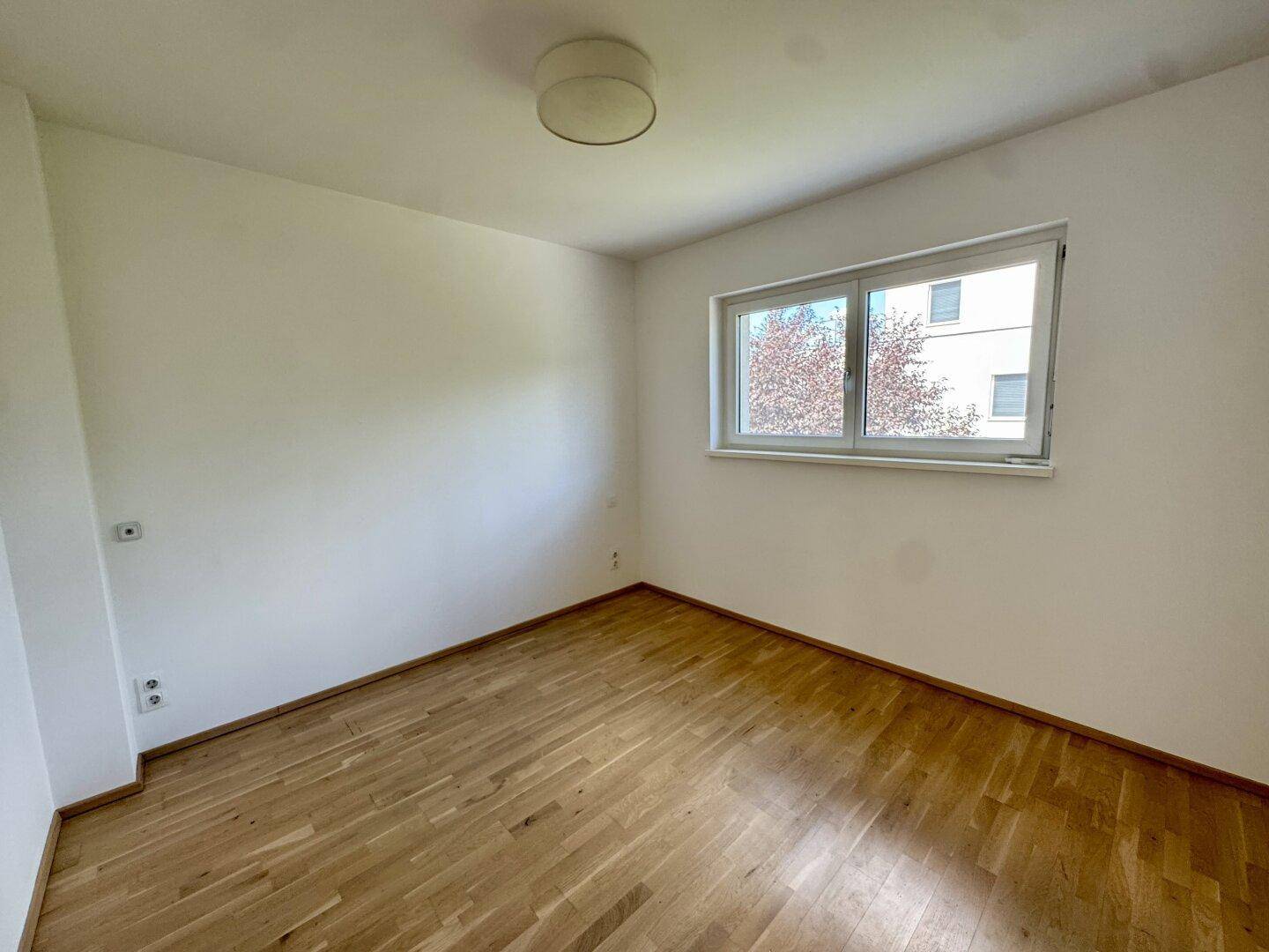 Leeres Zimmer mit Holzparkett und Fenster mit Blick ins Grüne.