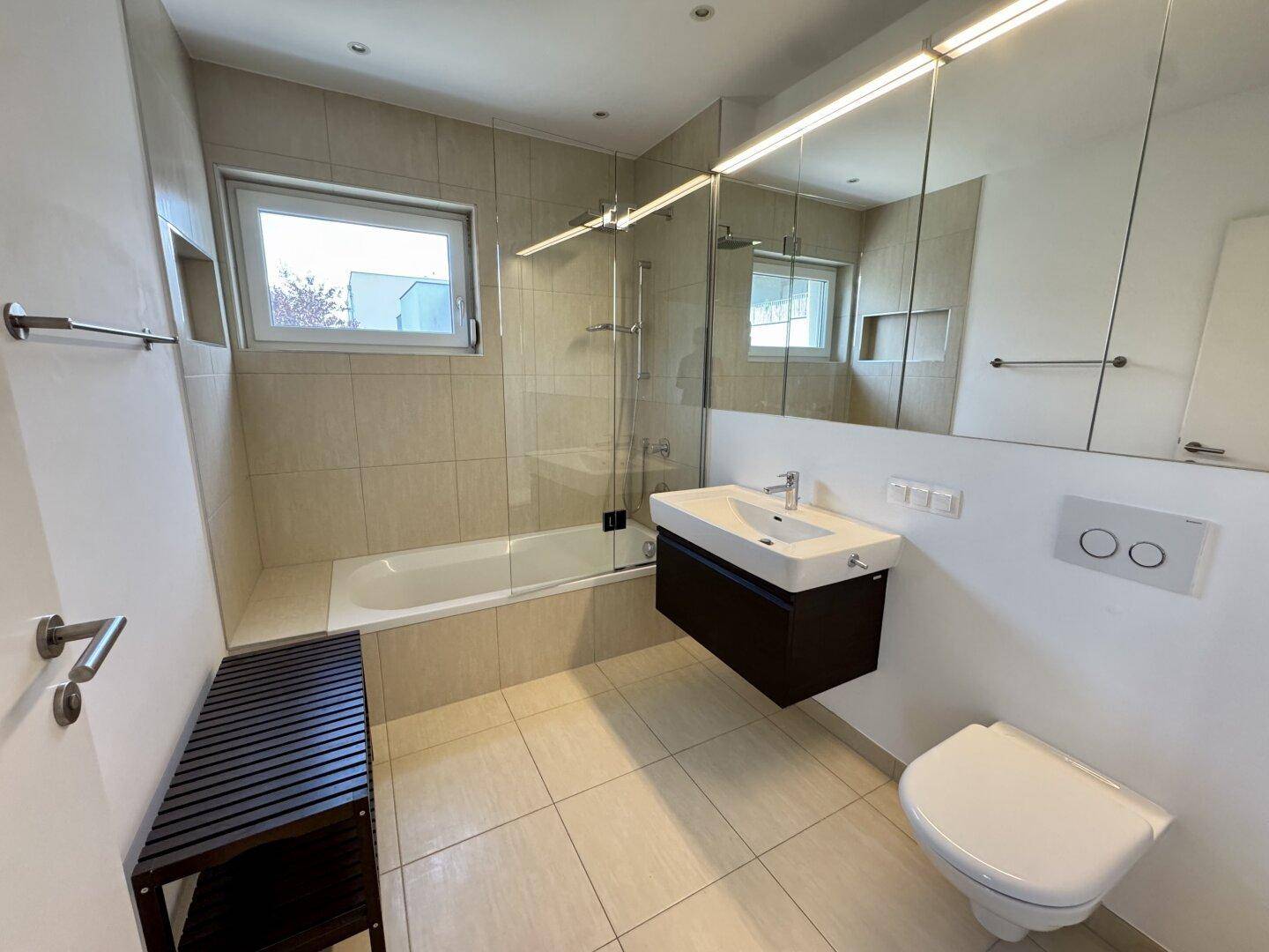 Modernes Badezimmer mit beigen Fliesen, Badewanne, schwebendem Waschtisch und Fenster.