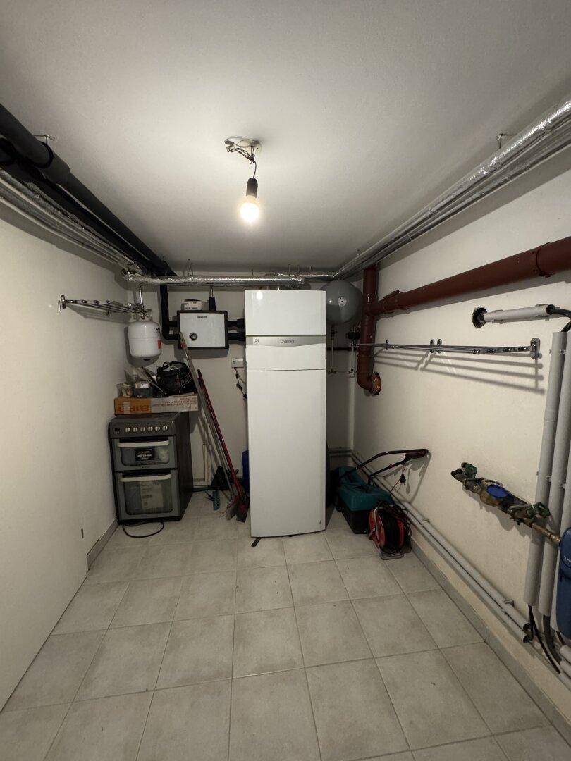 Kellerraum mit Stauraum, Kühlschrank und sichtbaren Versorgungsleitungen.