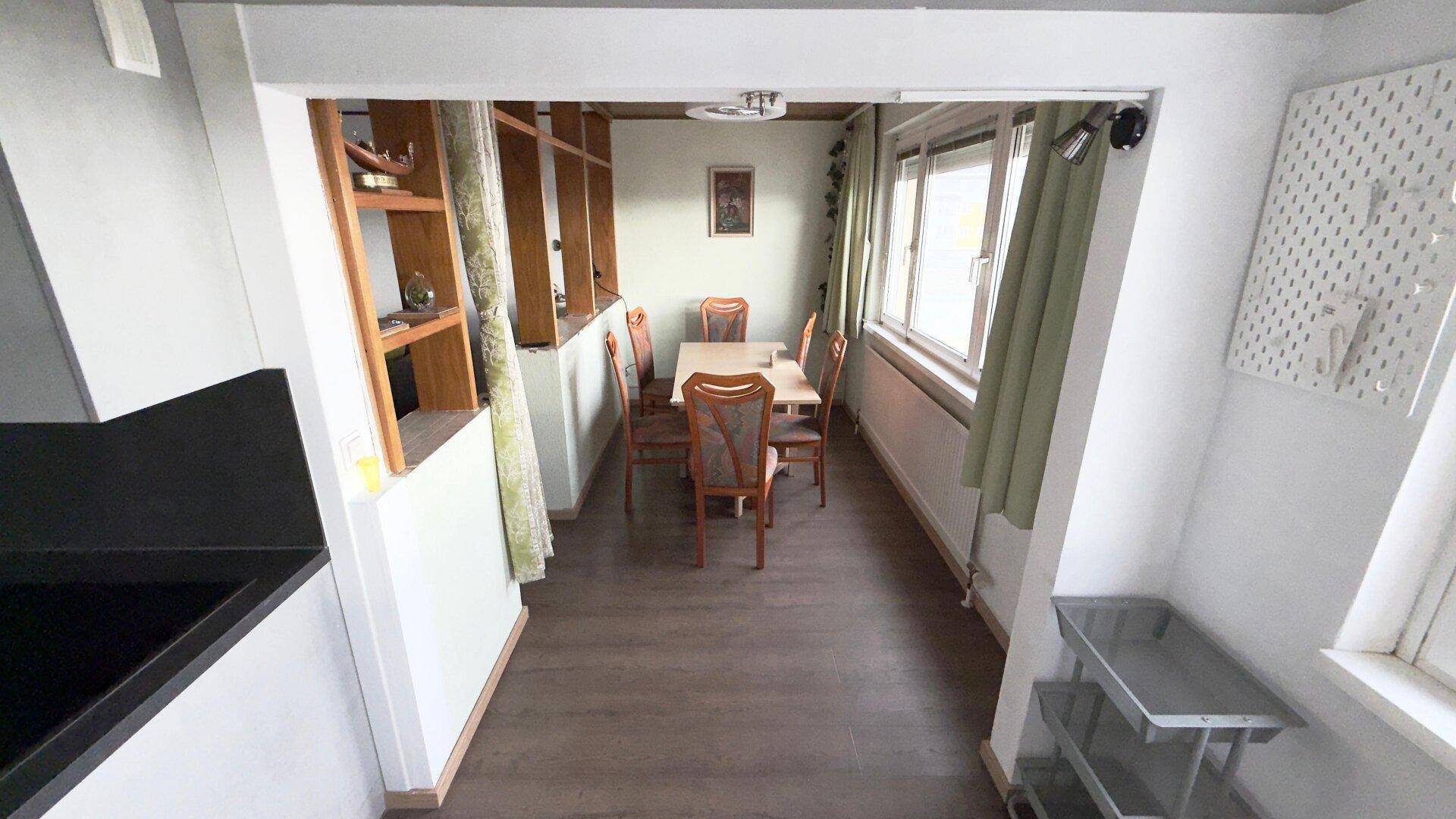 Heller Essbereich mit Holztisch und Stühlen, durch ein Fenster mit grünen Vorhängen natürlich belichtet.