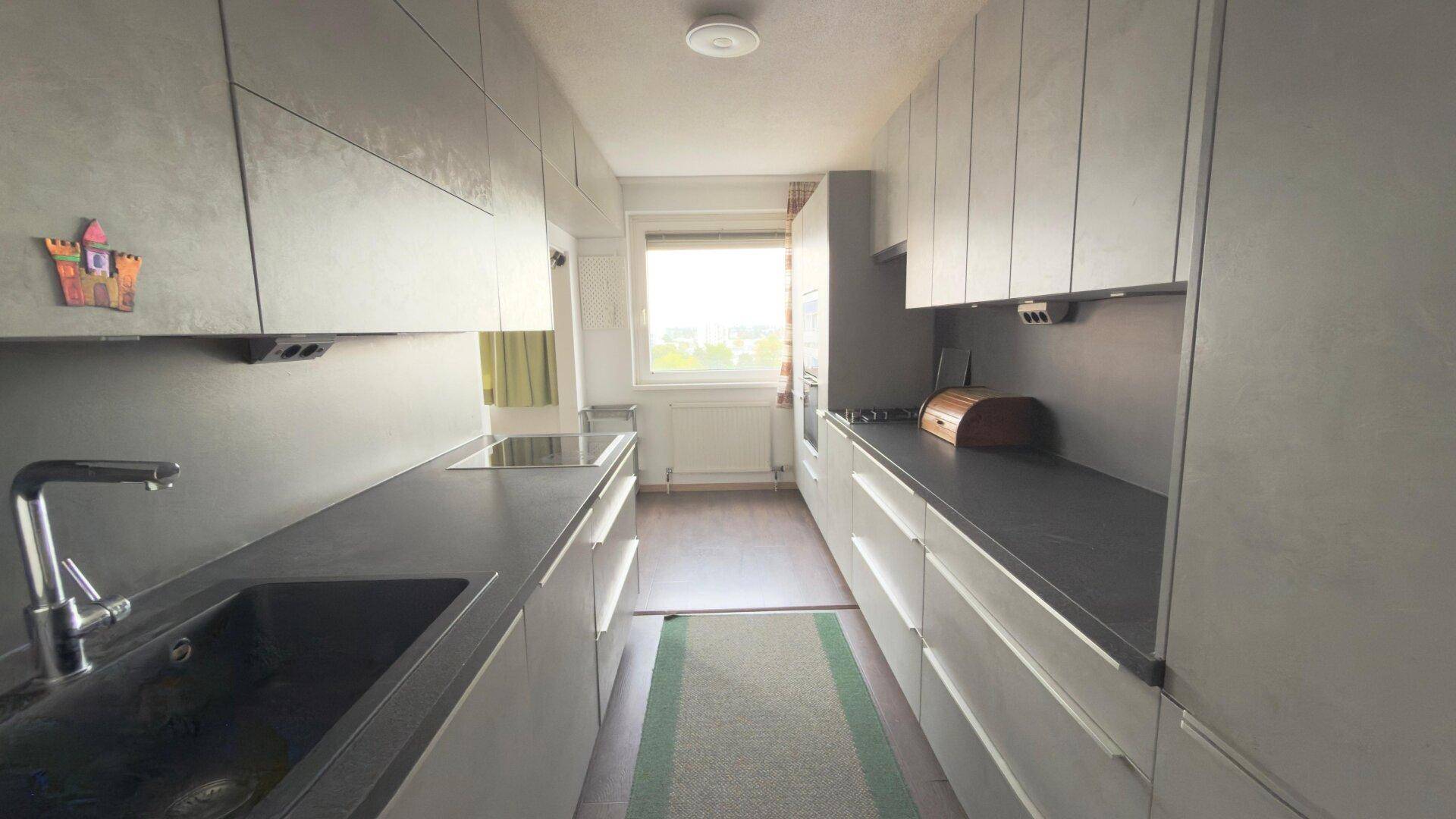 Moderne Küche in Grau mit Einbauschränken, dunklen Arbeitsplatten und Induktionskochfeld, hell durch das Fenster.