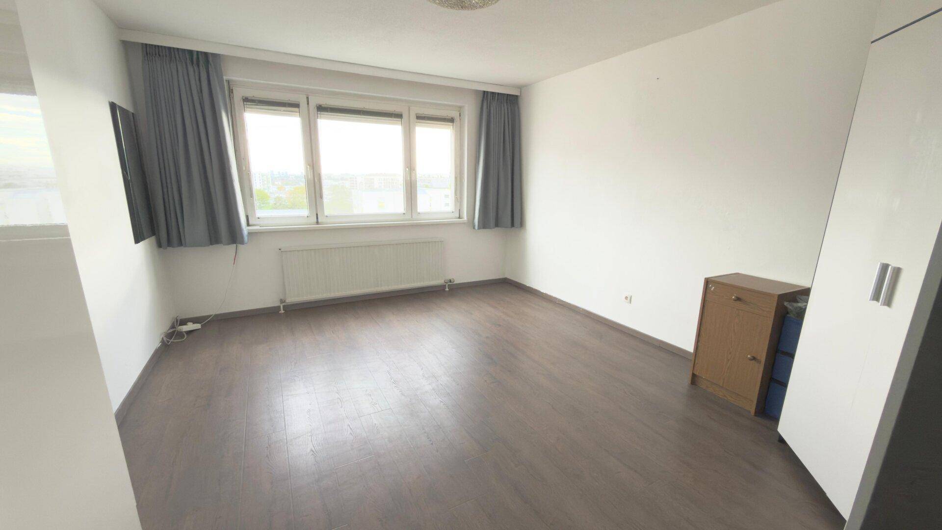 Helles Schlafzimmer mit großem Fenster, Heizkörper und dunklem Laminatboden, bereit zur individuellen Gestaltung.