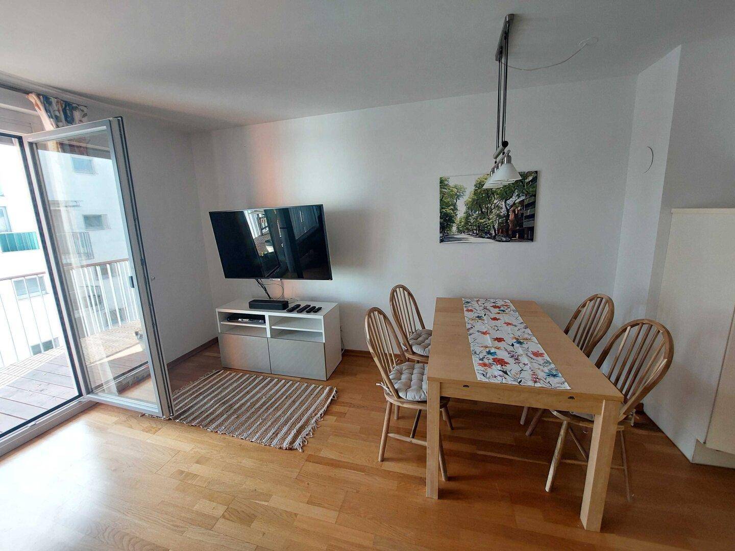 Heller Wohn- und Essbereich mit Holztisch, Fernseher und Zugang zu einem Balkon.