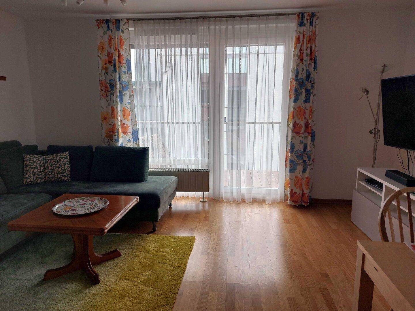 Gemütliches Wohnzimmer mit Ecksofa, Holztisch und großen Fenstertüren zum Balkon.