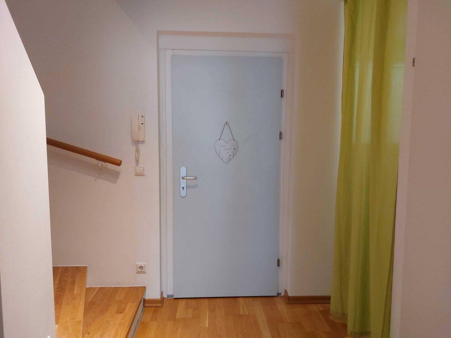 Heller Flur mit Eingangstür, Holztreppe und einer Gegensprechanlage.