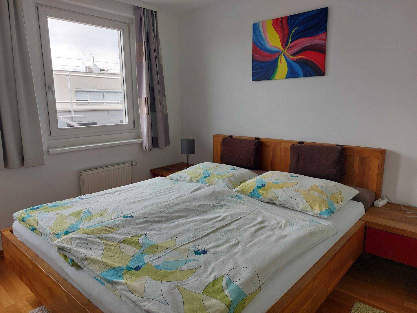 Schlafzimmer mit Doppelbett aus Holz, Nachttischen und einem farbenfrohen Kunstwerk an der Wand.