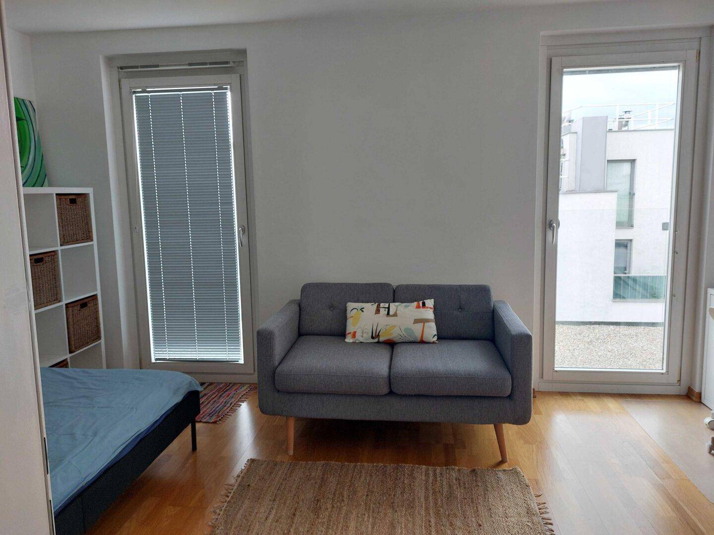 Heller Raum mit einem kleinen Sofa und großen Fenstern, die viel Tageslicht hereinlassen.