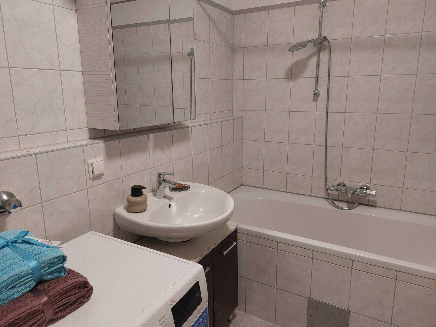 Gefliestes Badezimmer mit Badewanne, Waschbecken und Spiegelschrank, inklusive Waschmaschine.