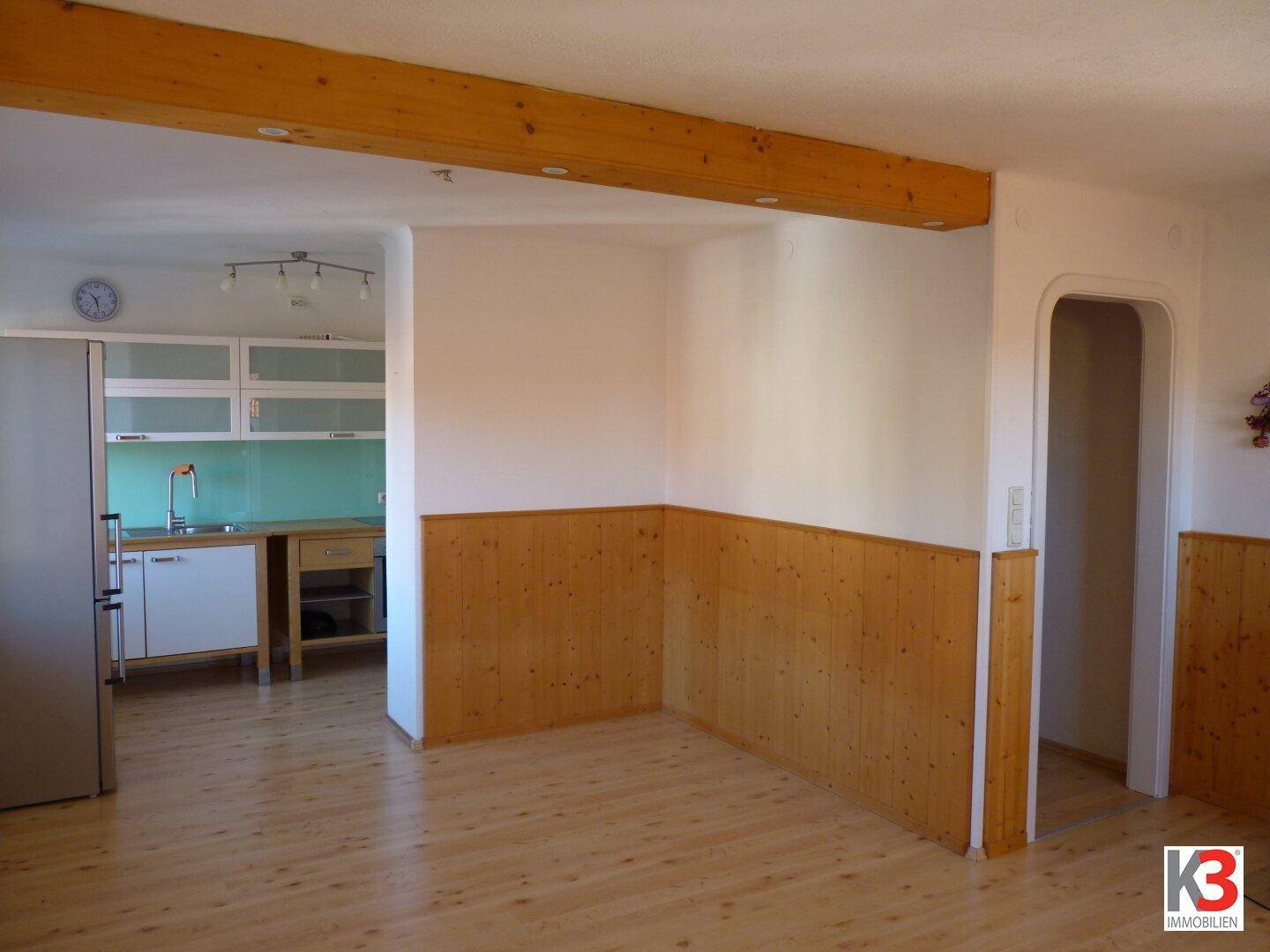 Offener Wohnbereich mit Holzboden, sichtbaren Holzbalken und angrenzender Küche mit hellen Schränken.