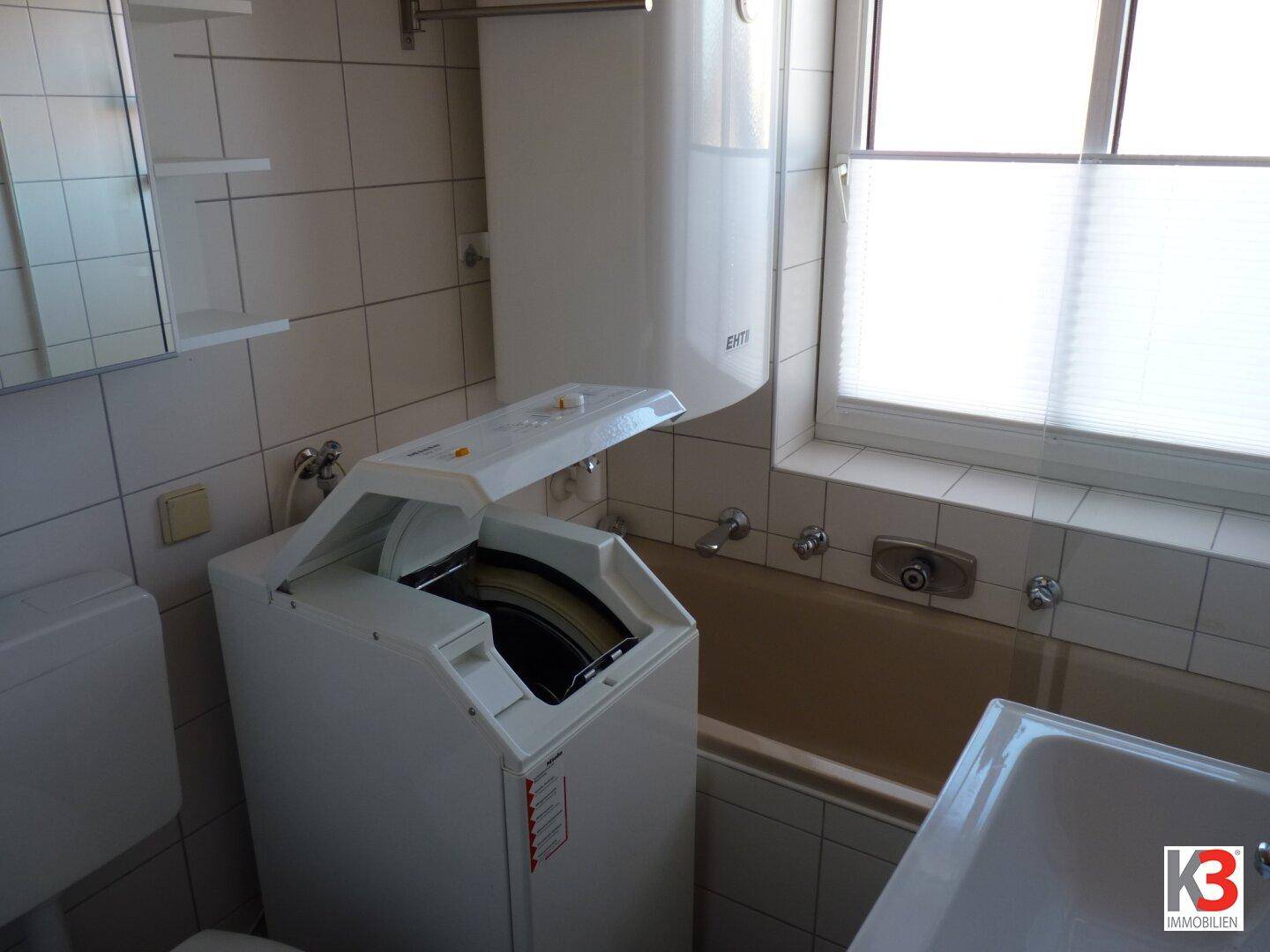 Funktionales Badezimmer mit Badewanne, Toilette und einer Waschmaschine, ausgestattet mit einem Fenster.