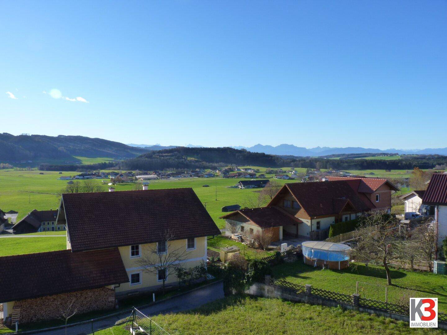 Weitläufige Landschaftsaufnahme mit mehreren Häusern, grünen Feldern und einer Bergkulisse im Hintergrund.