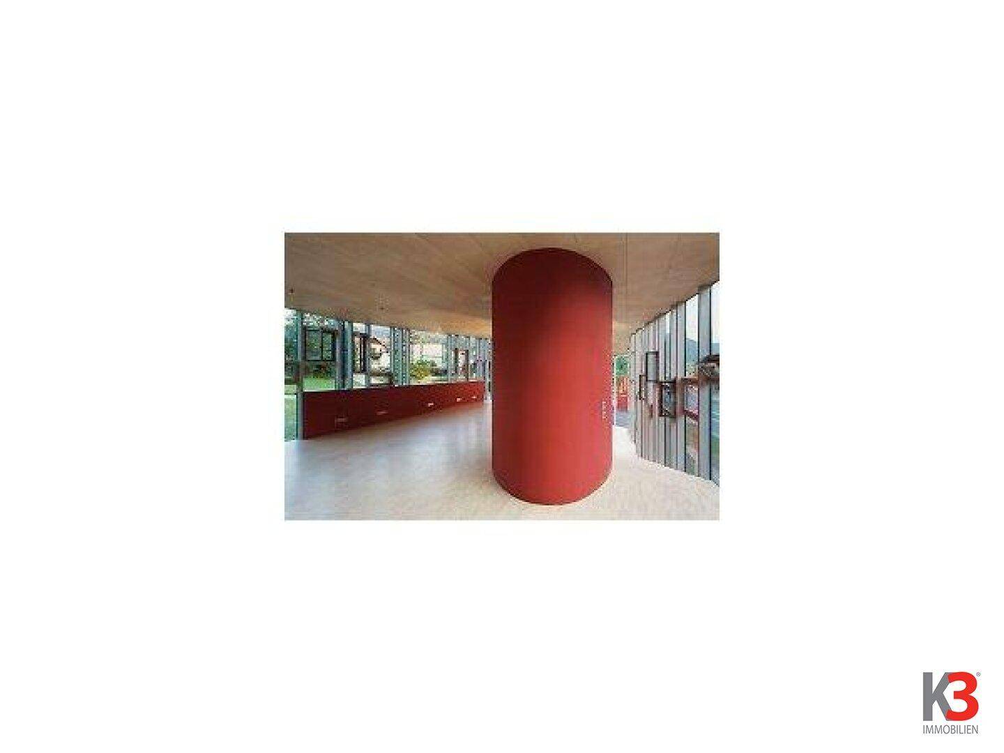 Innenraum mit markanter roter Säule, Holzdecke und großen Fensterfronten im Hintergrund.