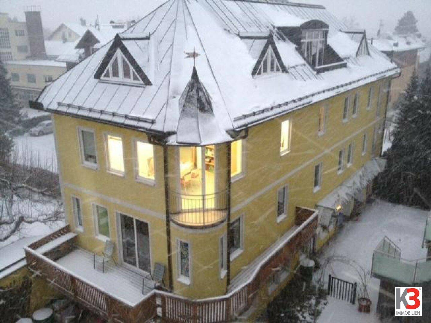 Gelbes Gebäude mit markantem Dach und Balkon, bedeckt mit Schnee, in winterlicher Umgebung.