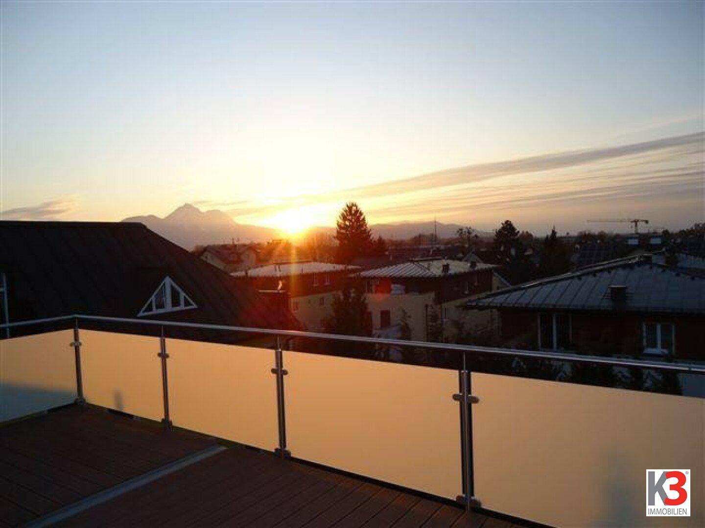 Großer Balkon mit Glasgeländer und Holzboden, bietet einen weiten Blick auf Berge bei Sonnenuntergang.