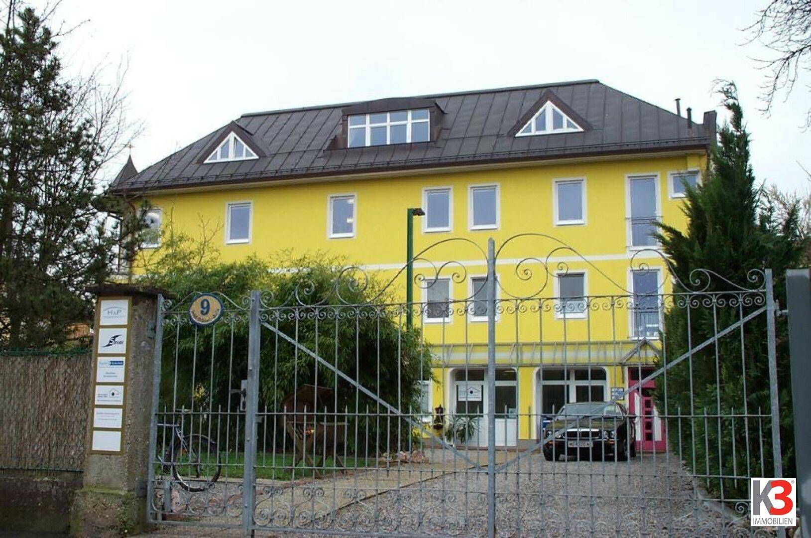 Gelbes Gebäude hinter einem schmiedeeisernen Tor mit Einfahrt und Büroeingangsbereich.