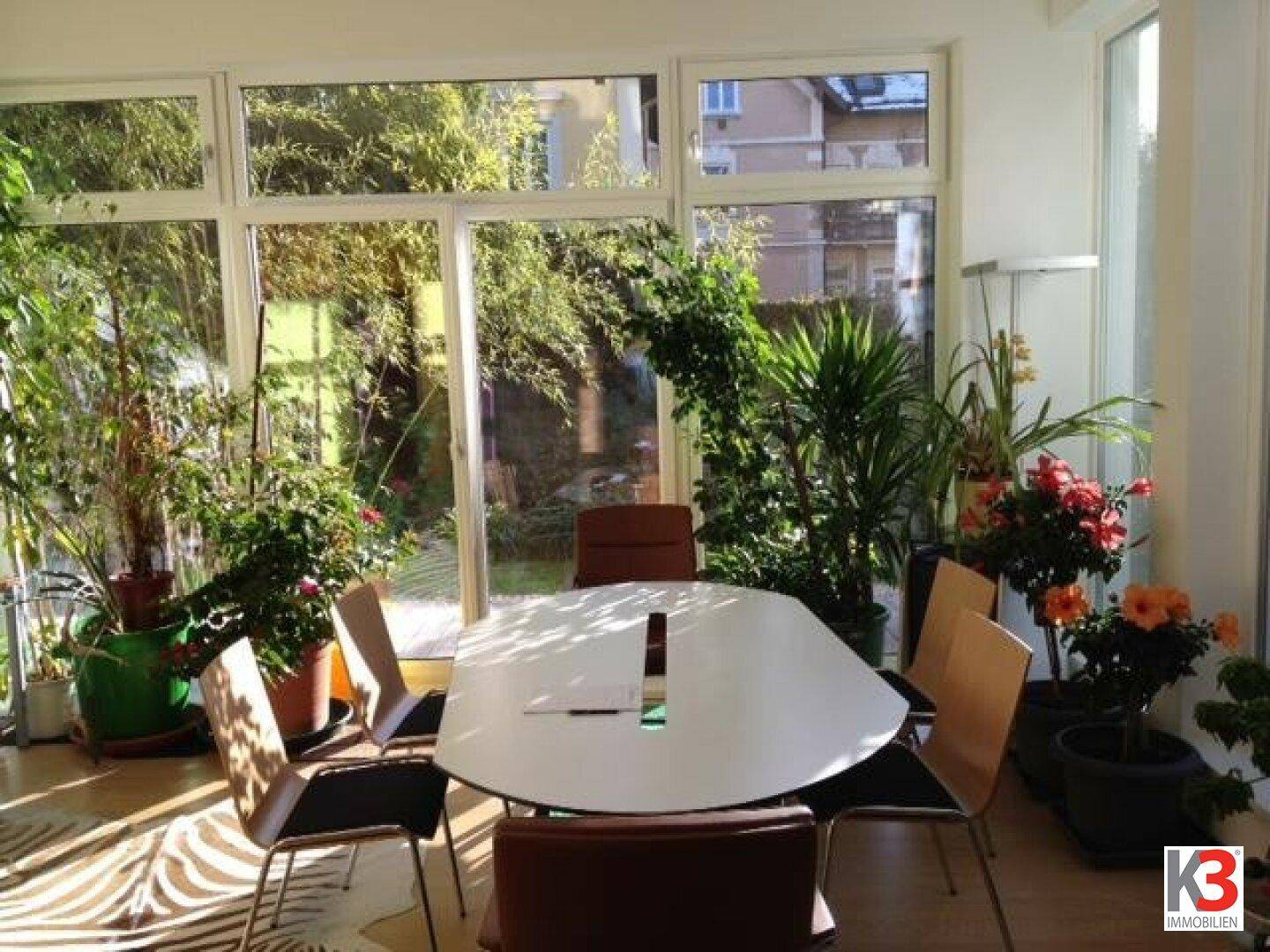 Heller Wintergarten mit großen Fenstern, vielen Pflanzen und einem modernen Esstisch mit Stühlen.