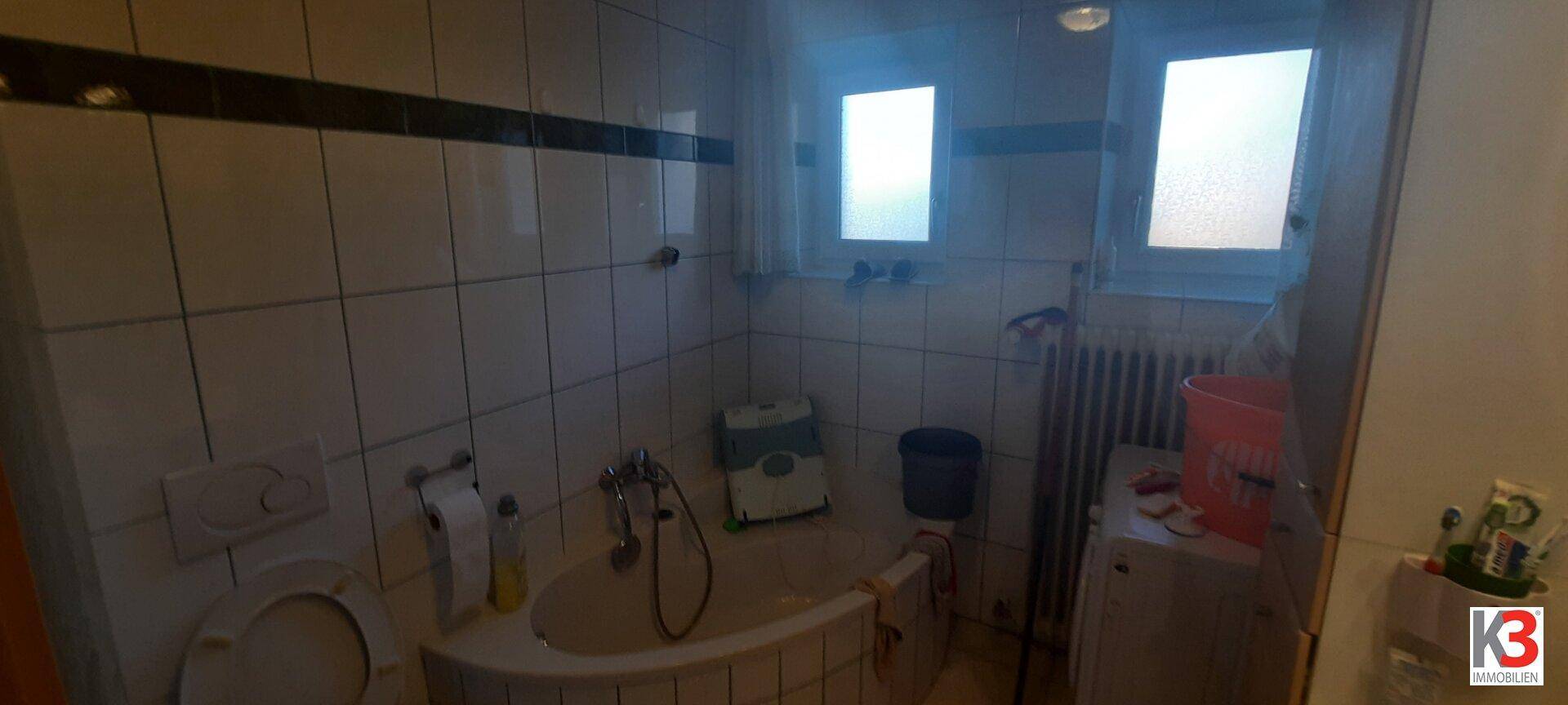 Badezimmer mit Badewanne, WC, weißen Fliesen und zwei Fenstern für natürliche Belüftung.