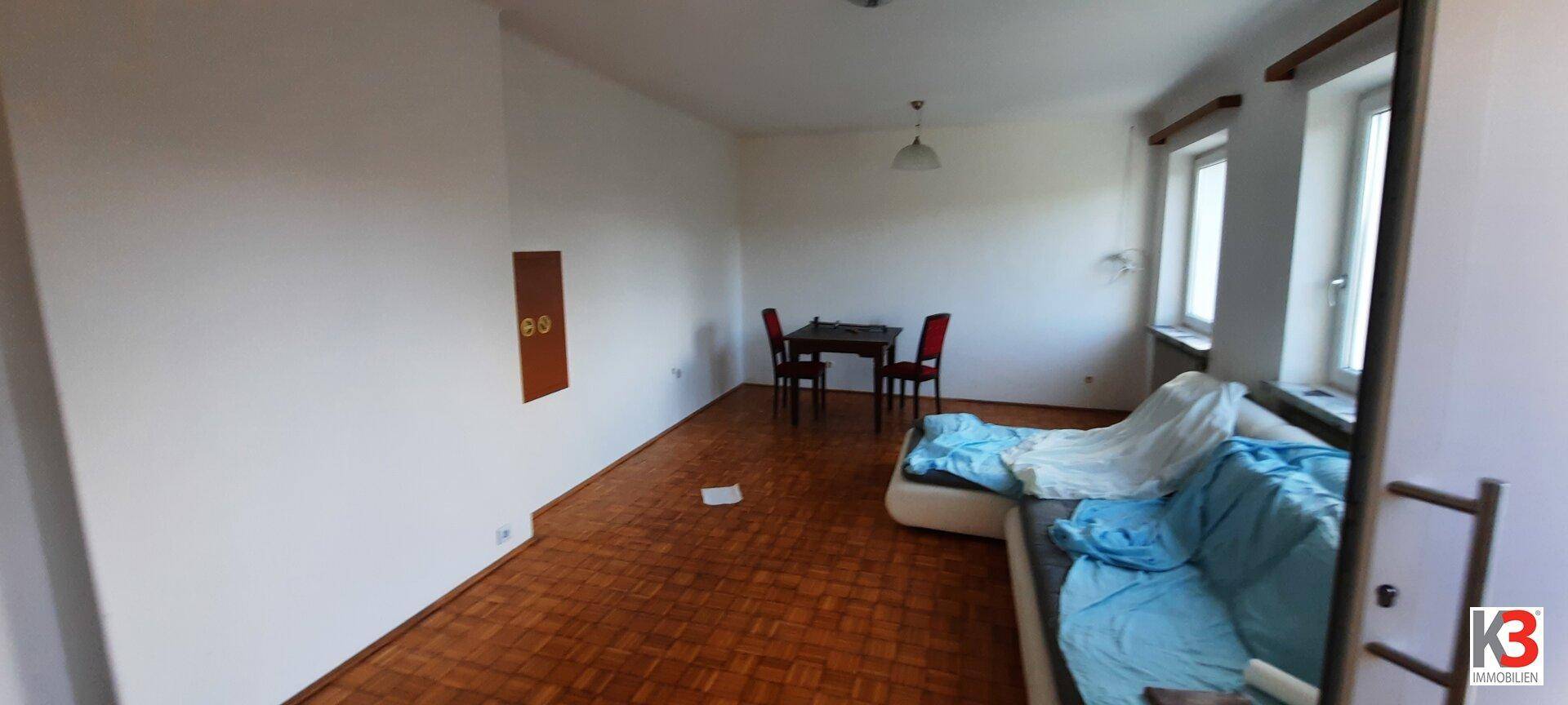 Heller Wohnraum mit Parkettboden, weißen Wänden, einem kleinen Esstisch und einem Sofa, der Renovierungsbedarf zeigt.