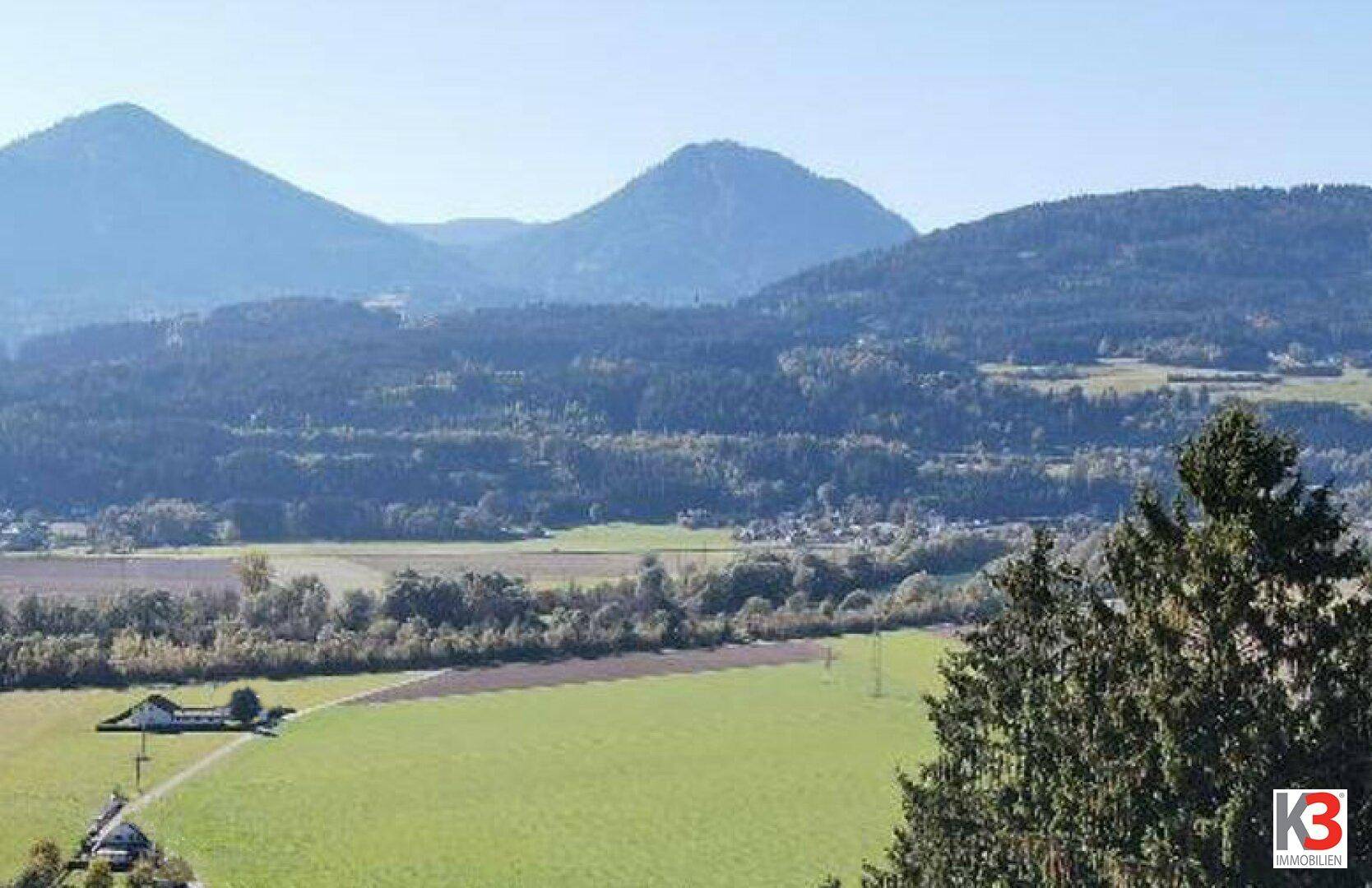 Weitläufiger Ausblick über grüne Felder und bewaldete Hügel mit vereinzelten Häusern in der Ferne.