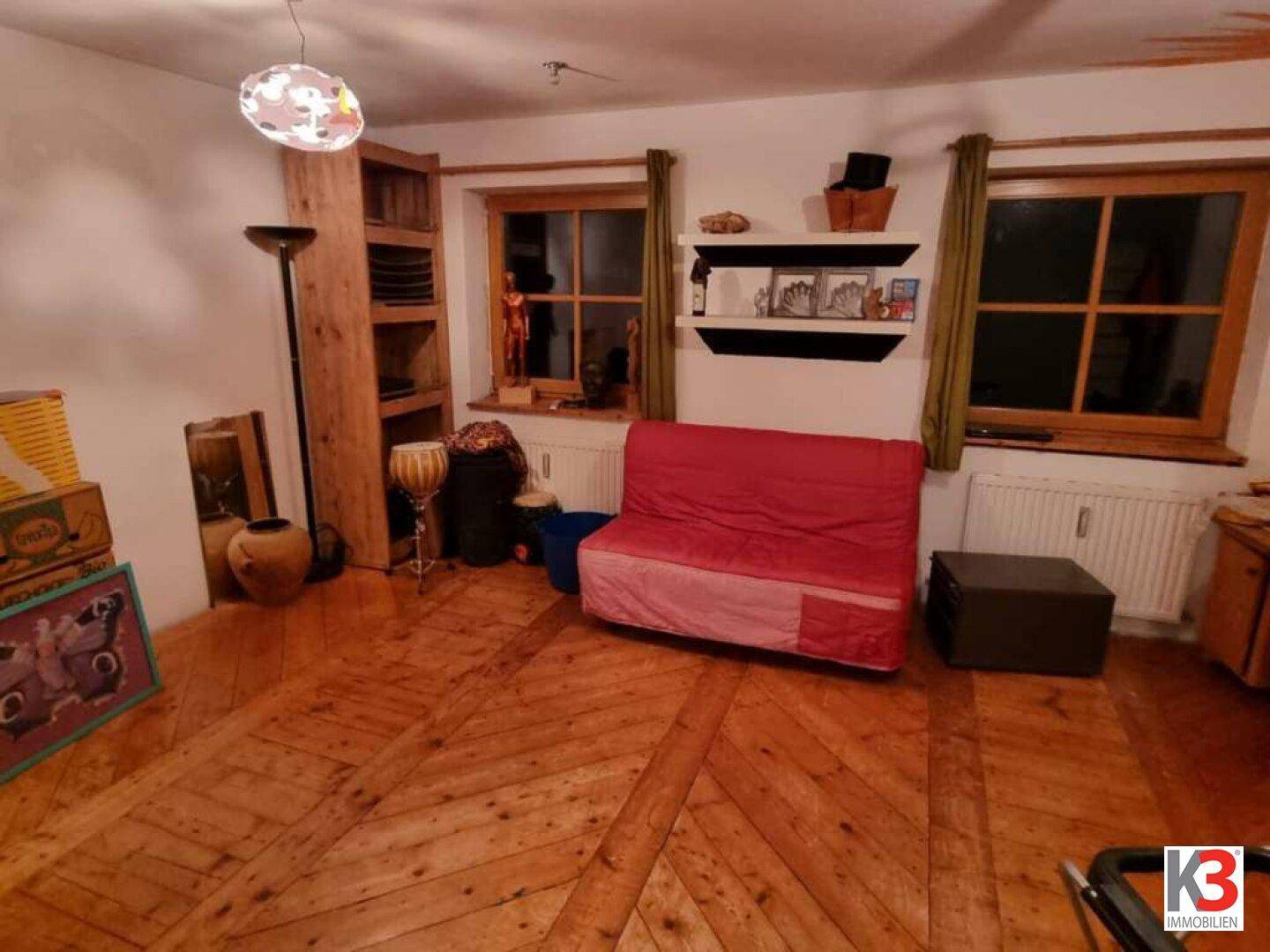 Helles Zimmer mit rotem Schlafsofa, einzigartigem Holzparkett und zwei Fenstern.