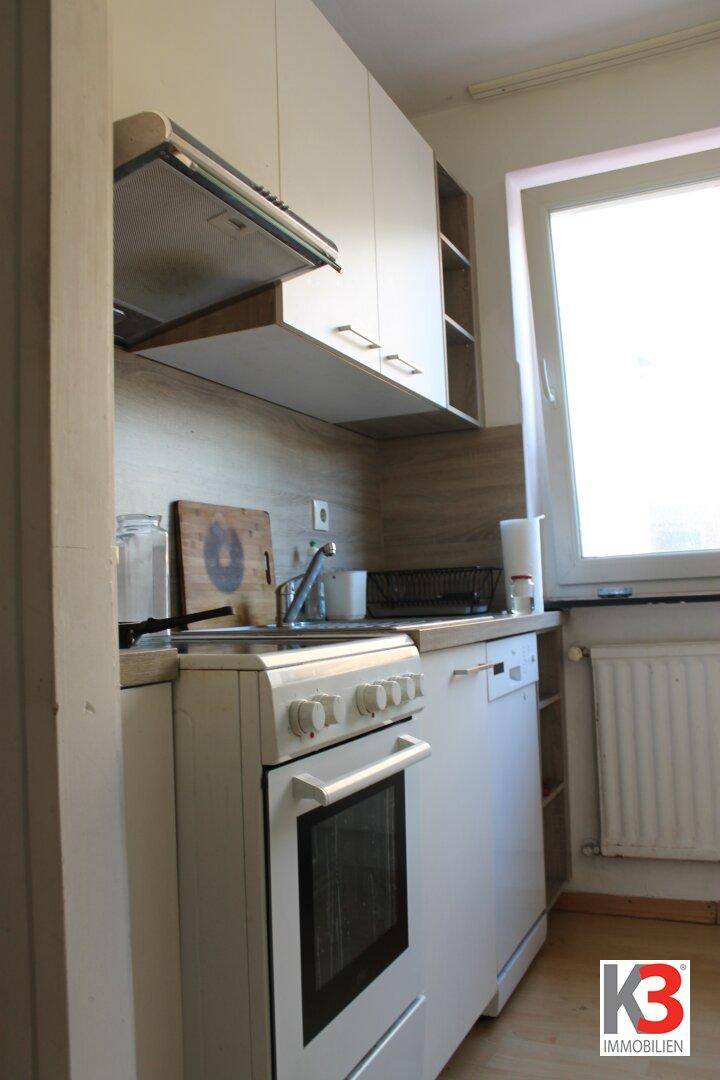Funktionale Küche mit weißen Schränken, Herd, Spülmaschine und einem Fenster für Tageslicht.