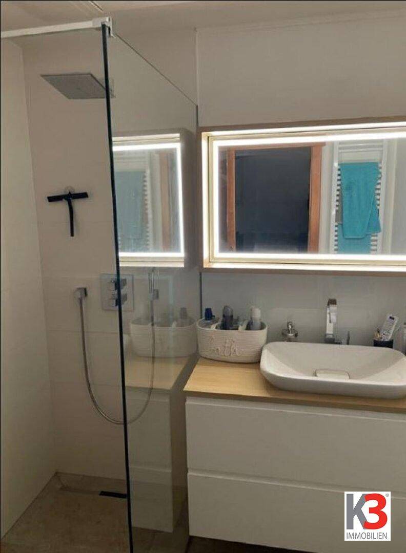 Modernes Badezimmer mit begehbarer Dusche, beleuchtetem Spiegel und Einzelwaschbecken.