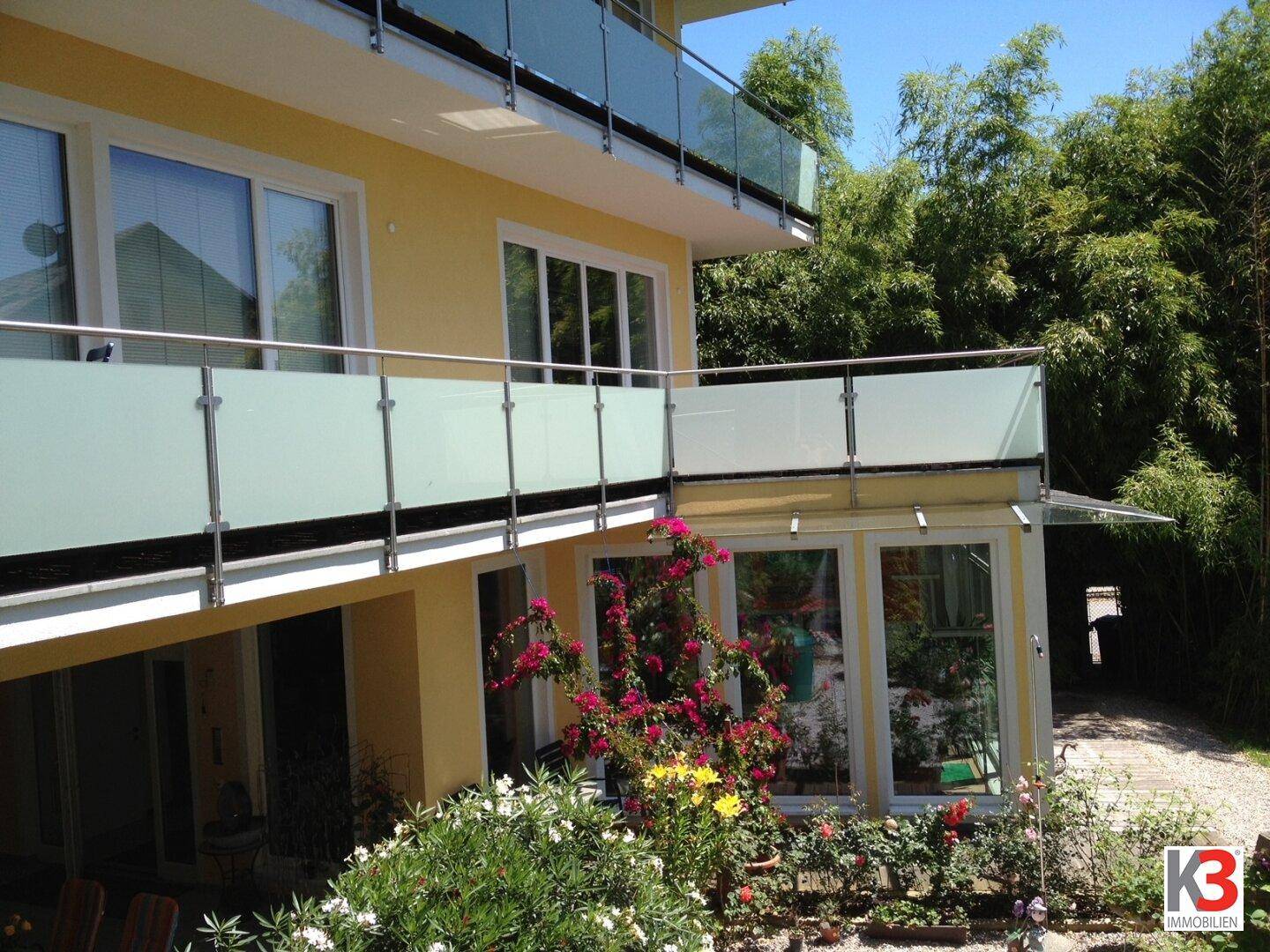 Außenansicht des Gebäudes mit blühenden Pflanzen und einem Balkon mit modernem Glasgeländer.