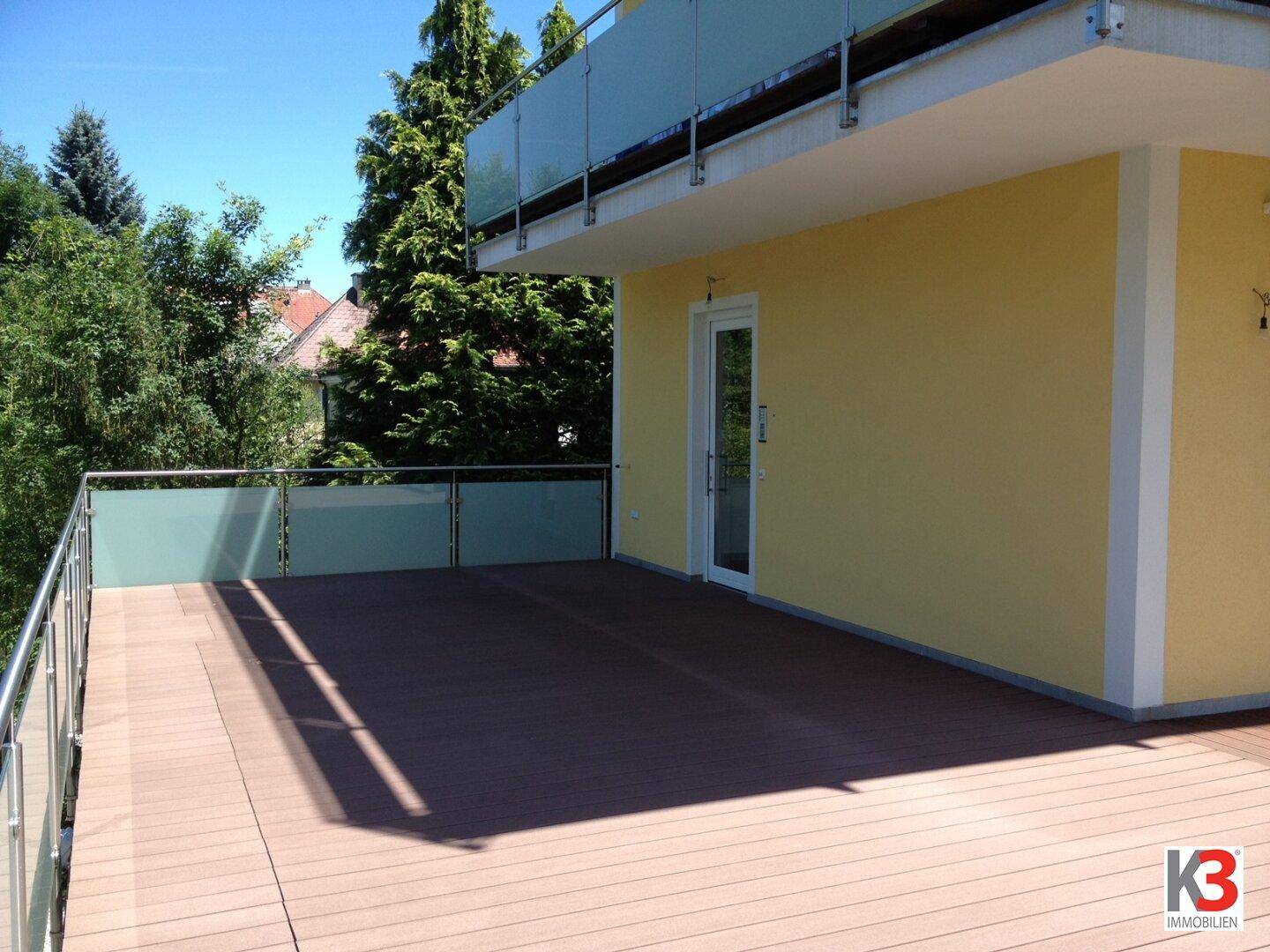Weitläufige Holzterrasse mit moderner Glasbrüstung und direktem Zugang zum Innenbereich des Hauses.