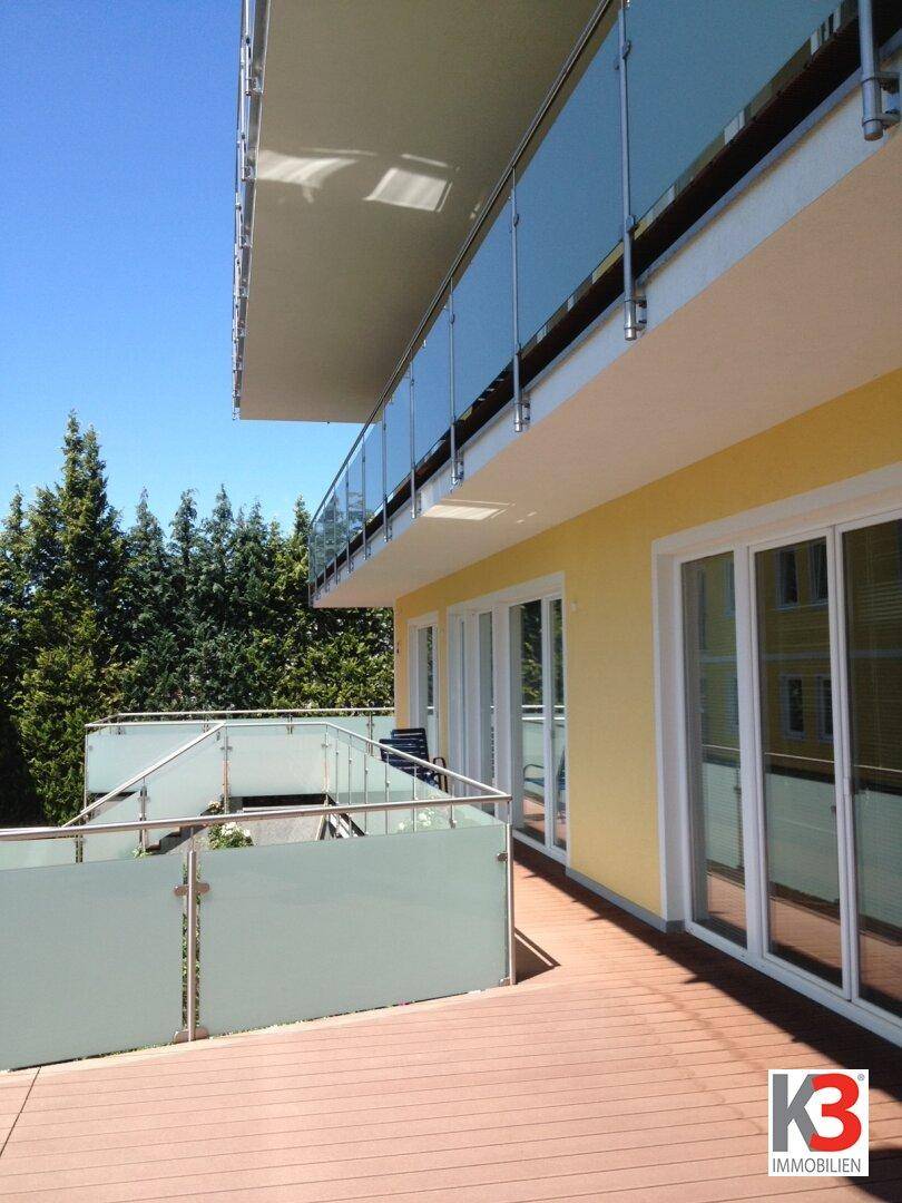 Moderne Holzterrasse mit Glasgeländer und weitem Blick auf die grüne Umgebung unter blauem Himmel.