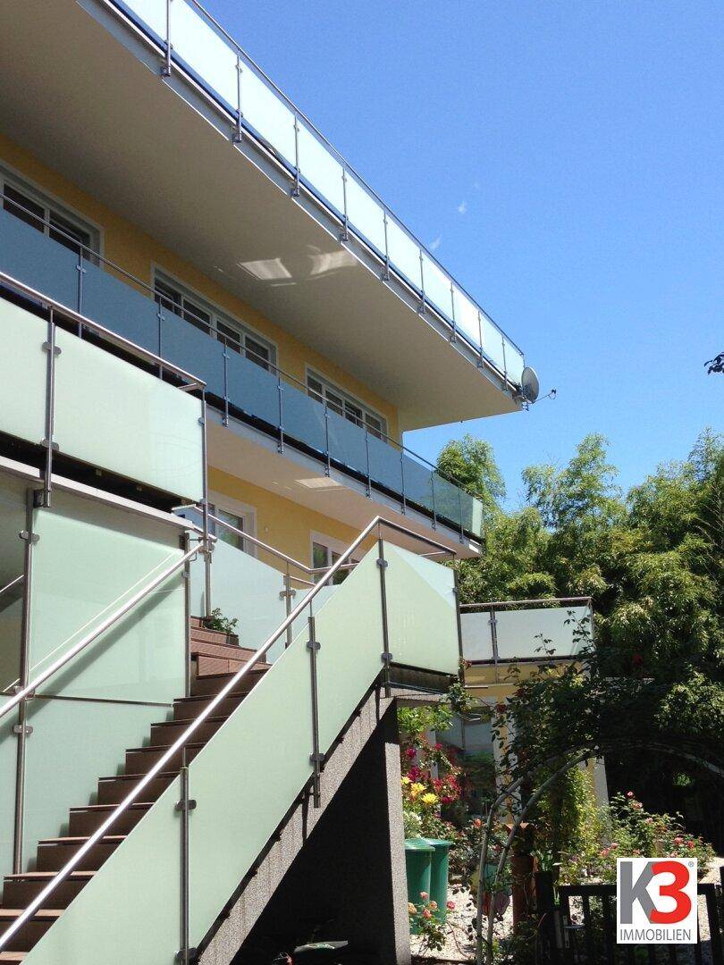 Elegante Außentreppe mit Glasgeländer und gelber Fassade, umgeben von viel Grün und blauem Himmel.
