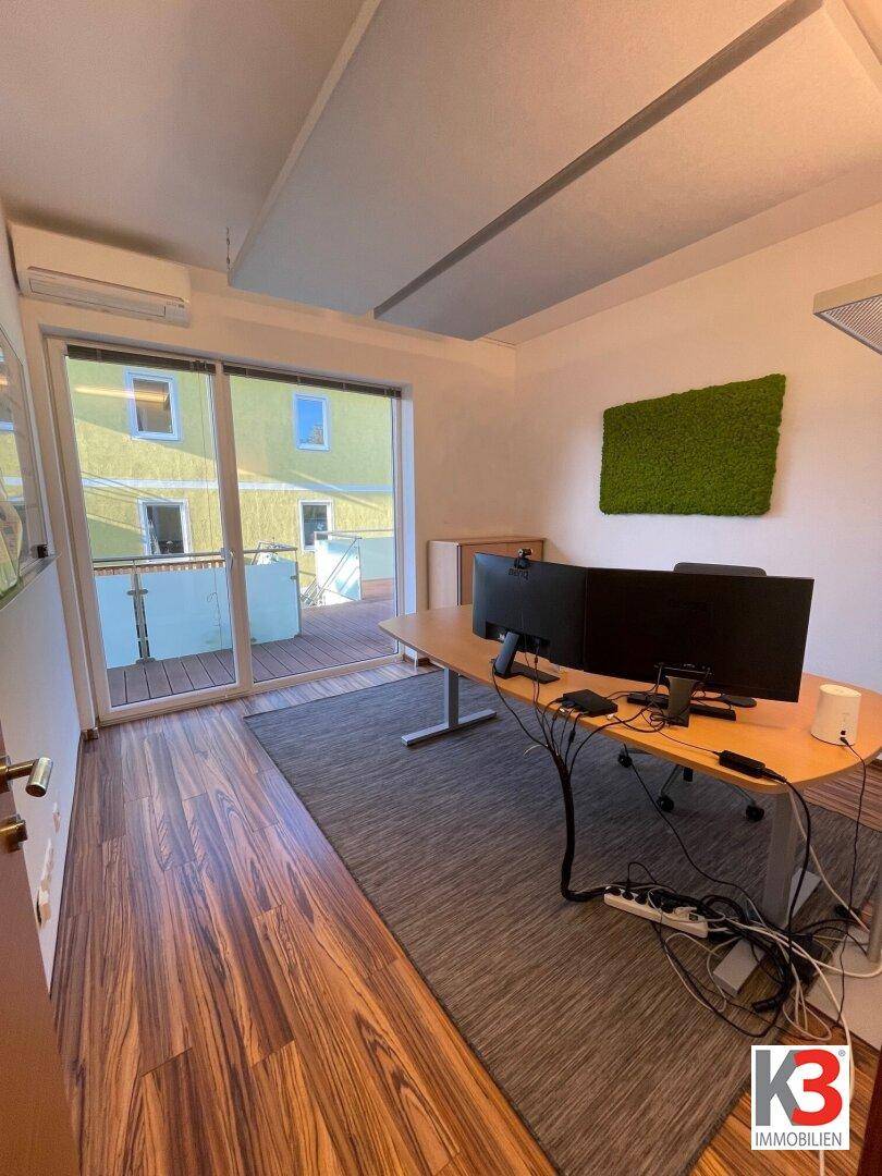 Modernes Büro mit großem Schreibtisch, zwei Monitoren und Zugang zum Balkon mit Blick ins Grüne.