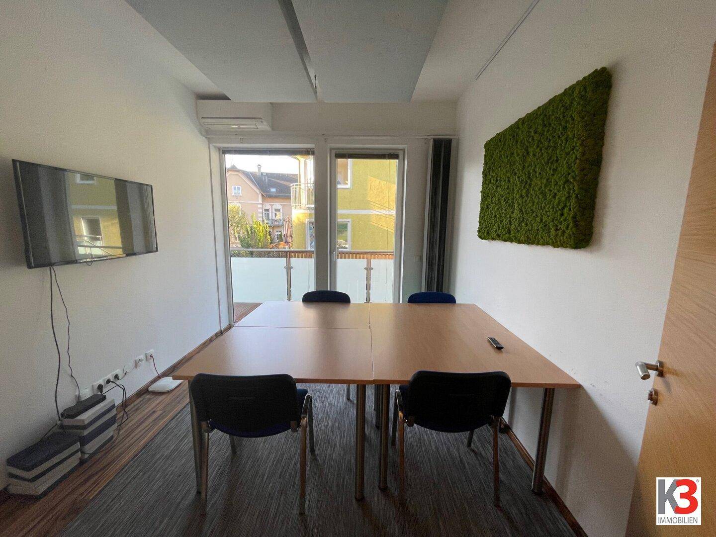 Besprechungsraum mit großem Tisch, Wandfernseher und Zugang zu einem Außenbereich mit viel Licht.