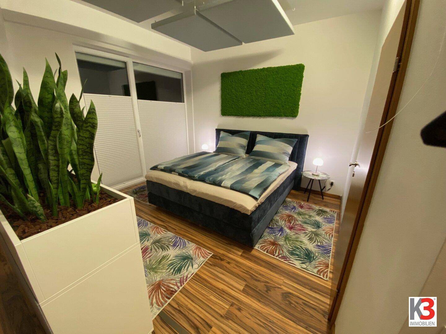 Helles Schlafzimmer mit Doppelbett, dekorativer Mooswand, Pflanzen und Fenster mit Jalousien.