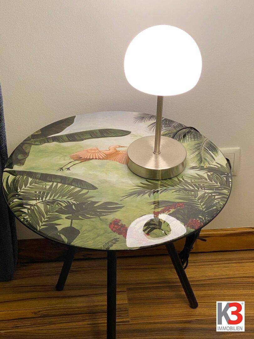 Detailaufnahme eines Nachttisches mit Lampe und tropischem Vogel- und Pflanzenmotiv.