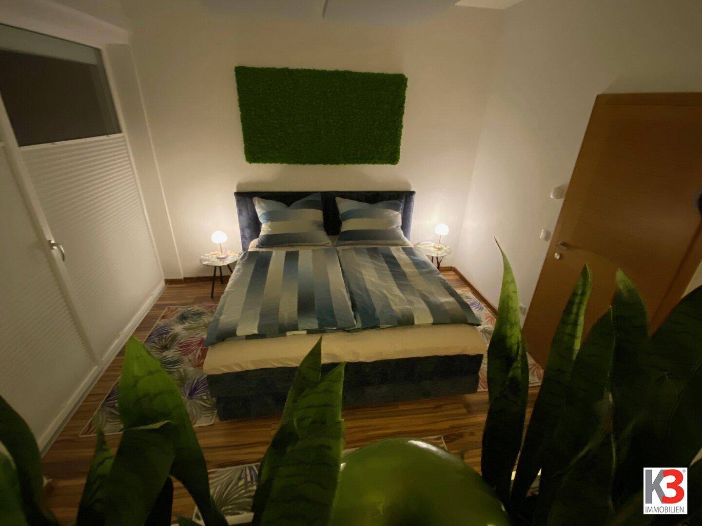 Schlafzimmer mit Doppelbett, Mooswanddekoration und Holzboden, hell beleuchtet.