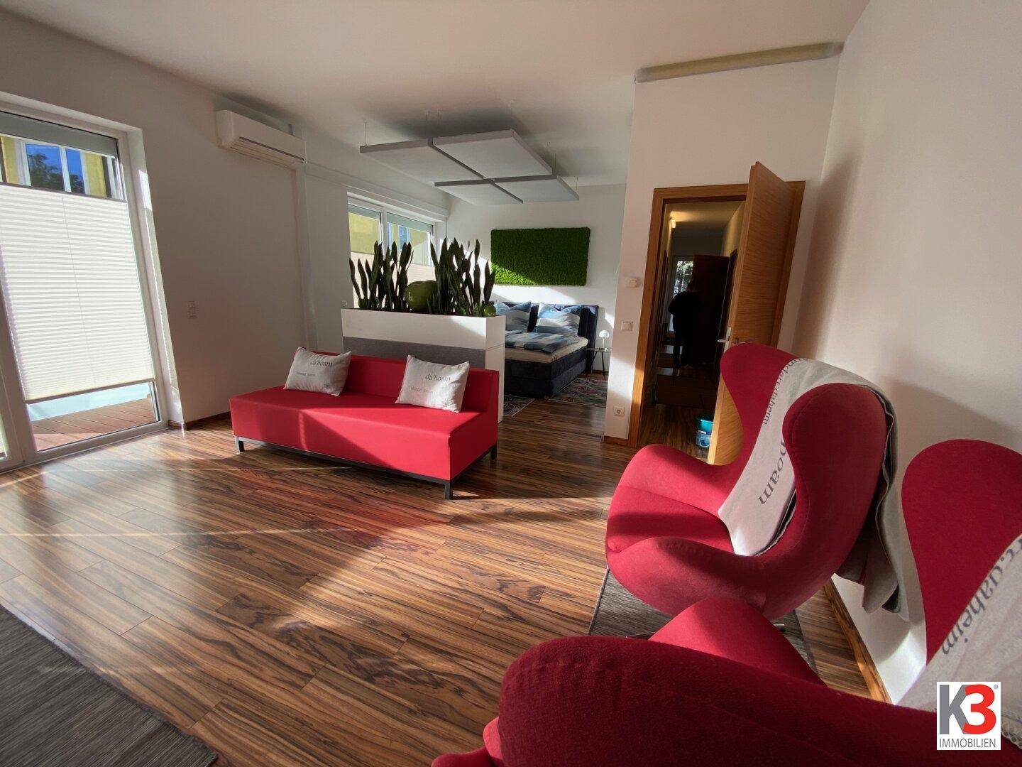 Geräumiger offener Wohnbereich mit rotem Sofa und Sesseln, verbunden mit dem Schlafbereich.