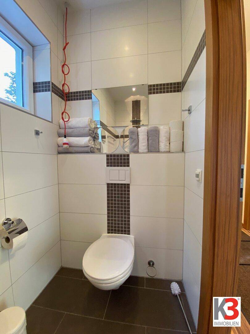 Modern gefliestes WC mit Wandtoilette, kleinem Fenster und dekorativen Handtüchern.