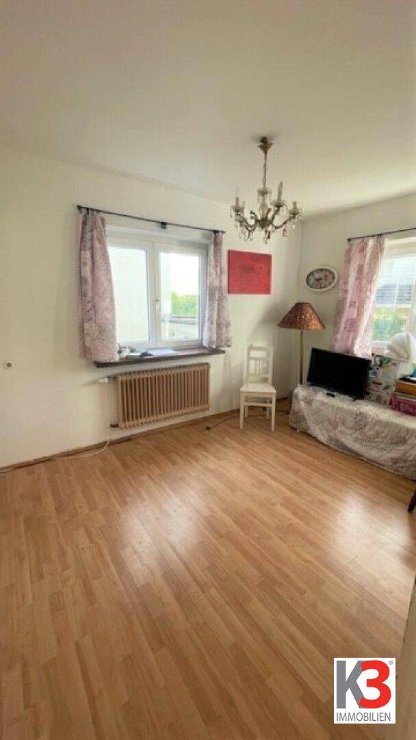 Helles Zimmer mit Holzboden, Fenster und einem klassischen Kronleuchter an der Decke.
