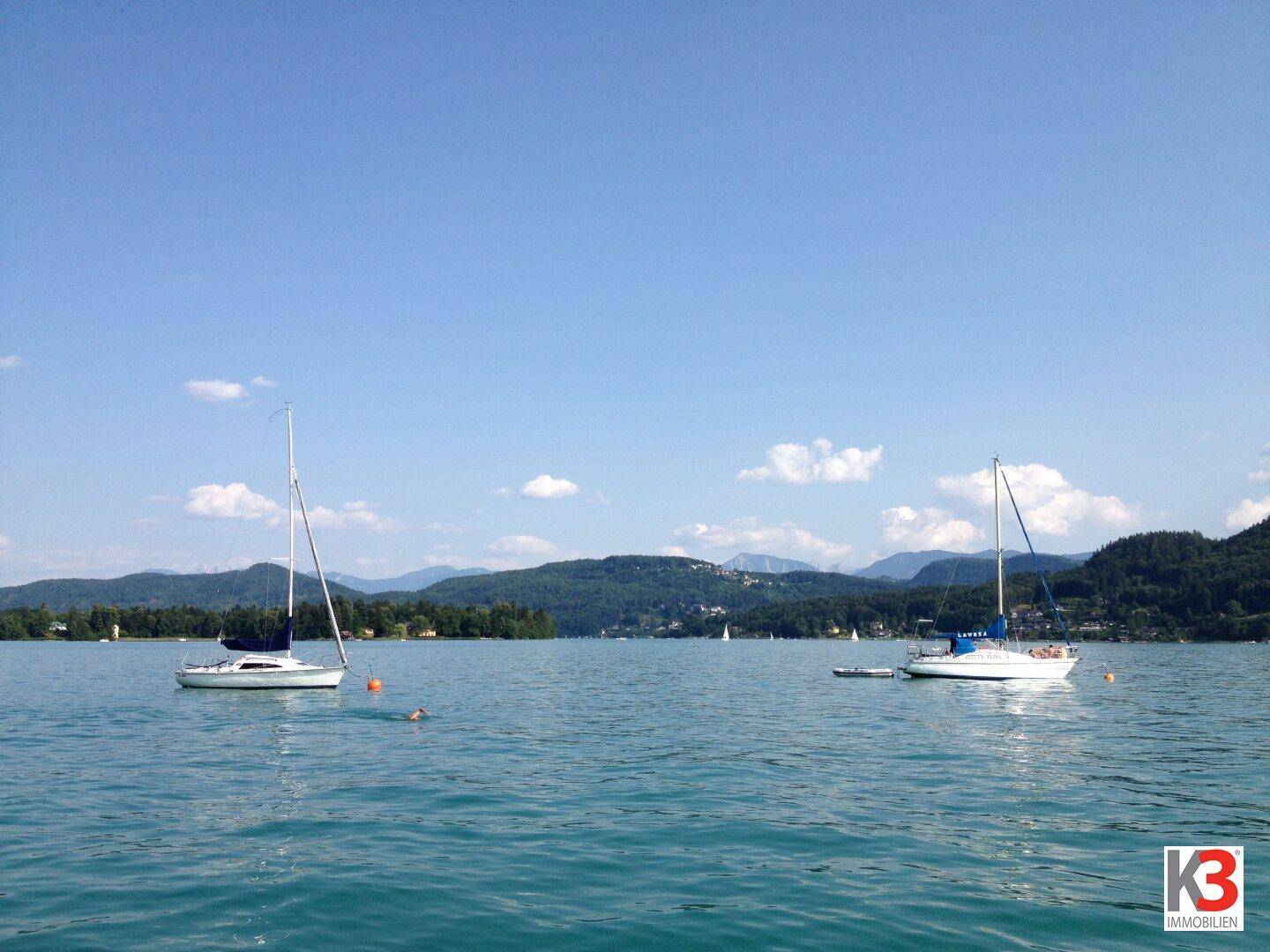 Ruhige Seelandschaft mit zwei Segelbooten auf dem Wasser und bewaldeten Bergen im Hintergrund unter blauem Himmel.
