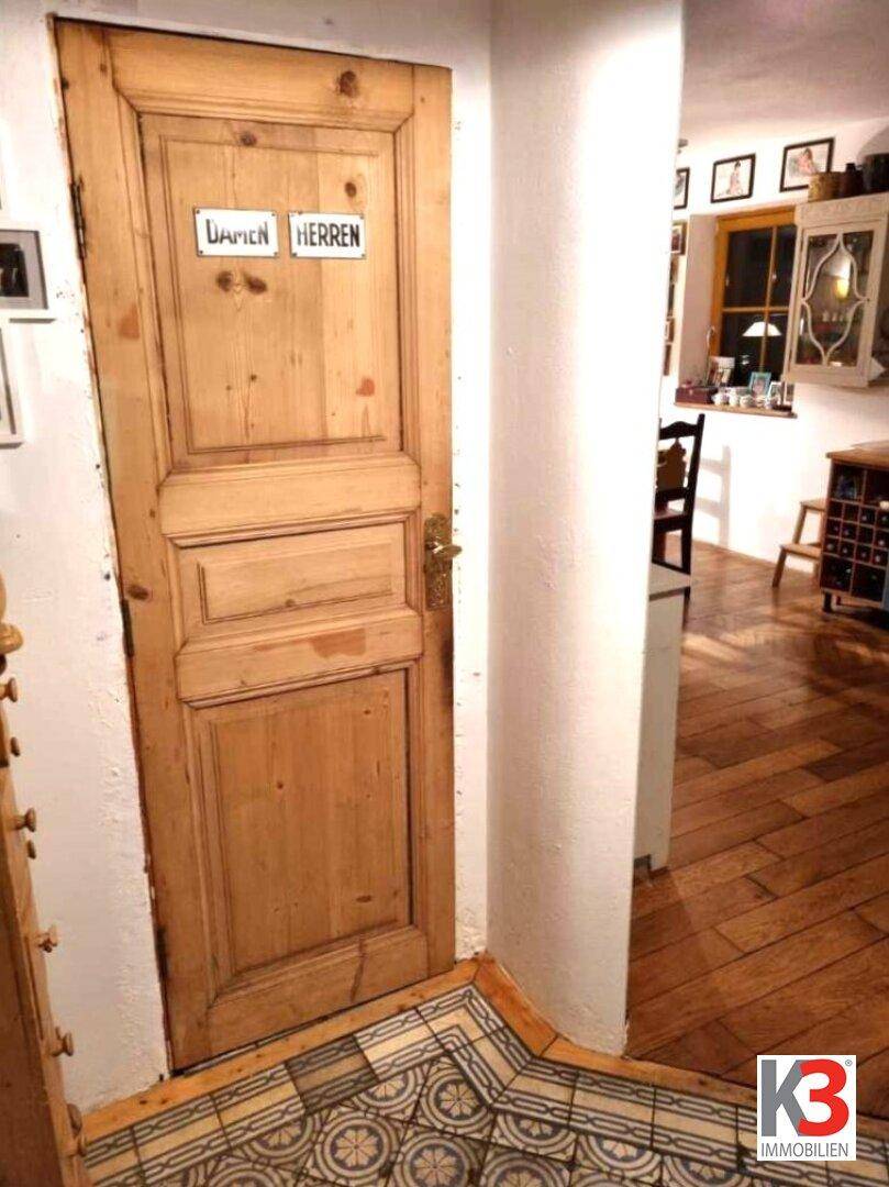 Holztür mit Beschriftung "Damen Herren" und gemustertem Fliesenboden im Eingangsbereich.
