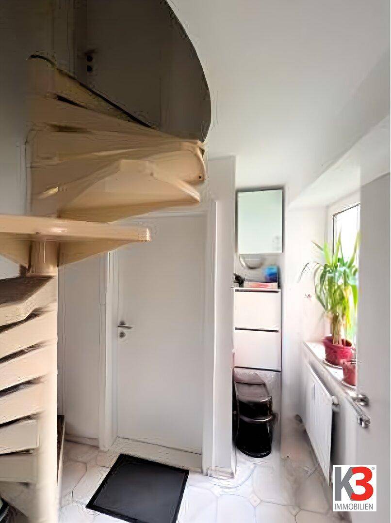 Heller Flur mit eleganter Holz-Wendeltreppe, weißer Tür und Fenster mit Grünpflanze.