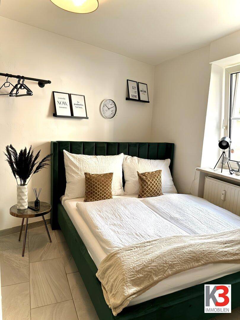Gemütliches Schlafzimmer mit grünem Polsterbett, Nachttisch und Fenster für natürliches Licht.