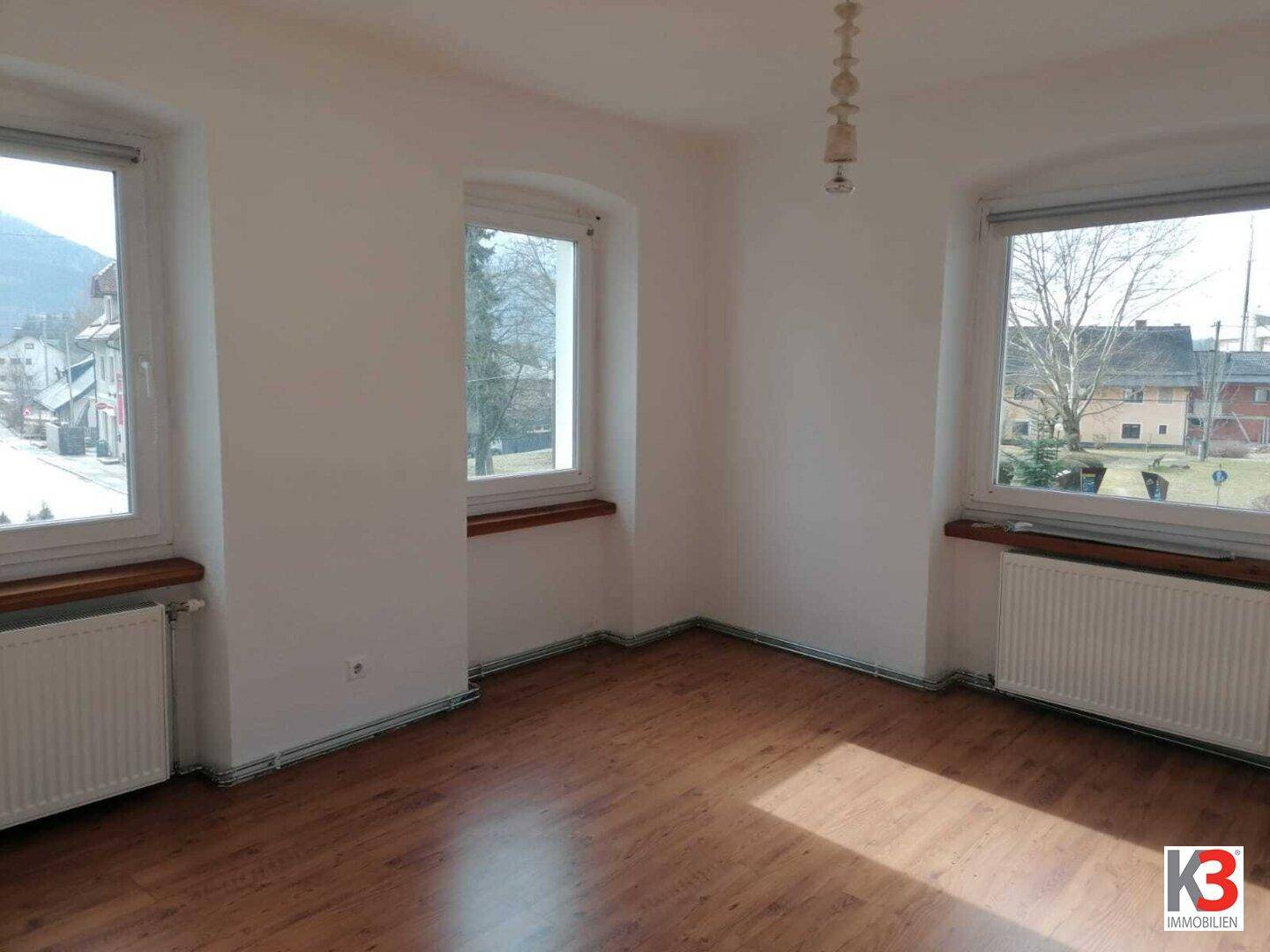 Helles, leeres Zimmer mit Holzboden, großen Fenstern und Blick auf die Umgebung.