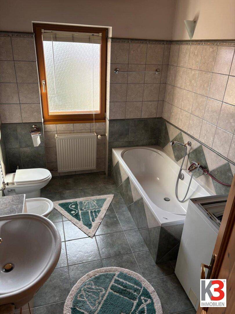 Badezimmer mit grünen und grauen Fliesen, Badewanne, Bidet und Fenster für natürliche Belüftung.