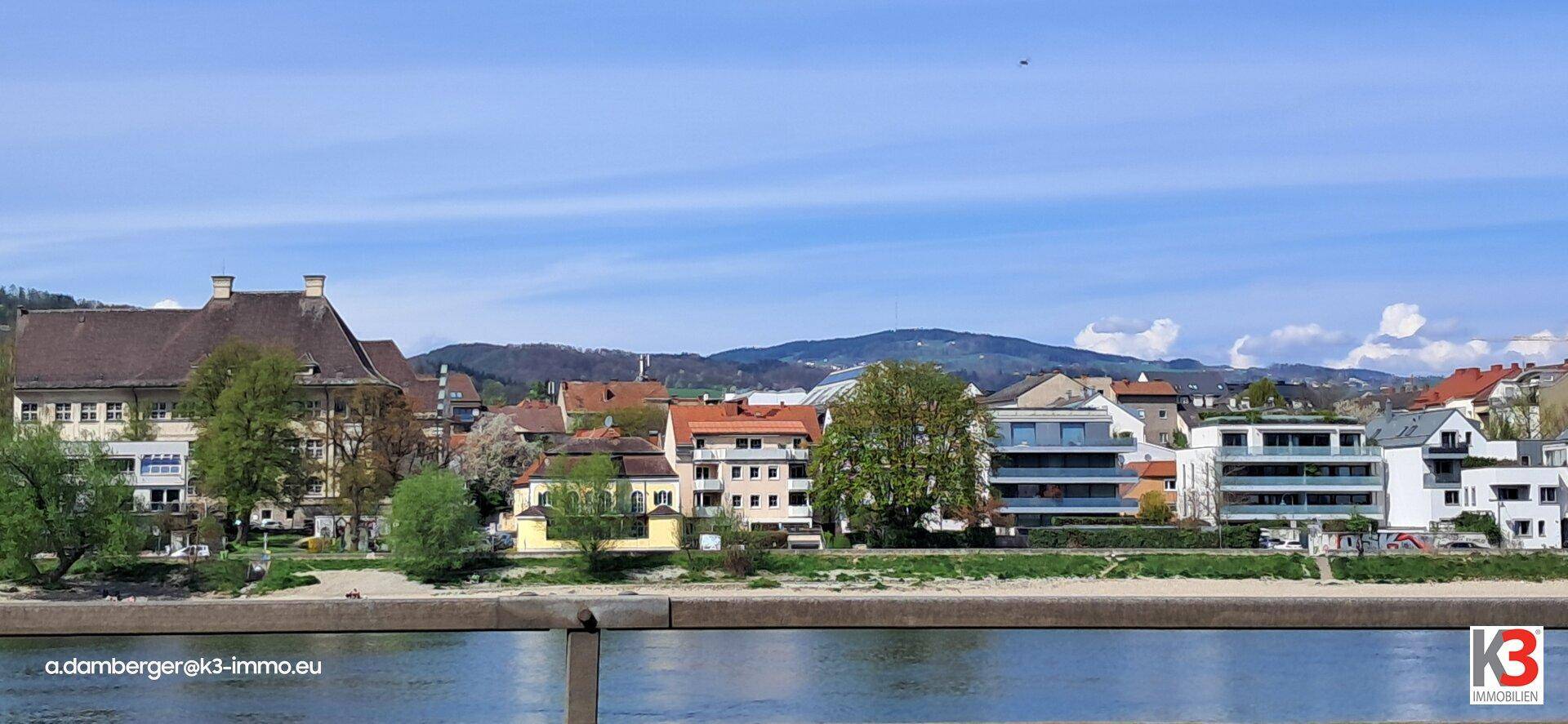 Panoramablick auf einen Fluss mit Gebäuden am Ufer und grünen Hügeln im Hintergrund.