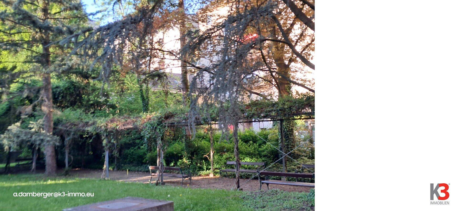 Üppiger Garten mit alten Bäumen, Sitzbänken und einer bewachsenen Pergola.